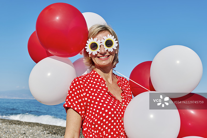 穿着红色波点裙的金发女人在阳光明媚的海滩上，手里拿着气球和搞笑眼镜。图片素材