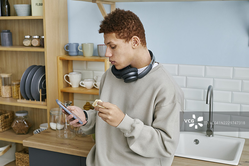 青少年在厨房吃早餐时使用手机，脖子上挂着耳机图片素材