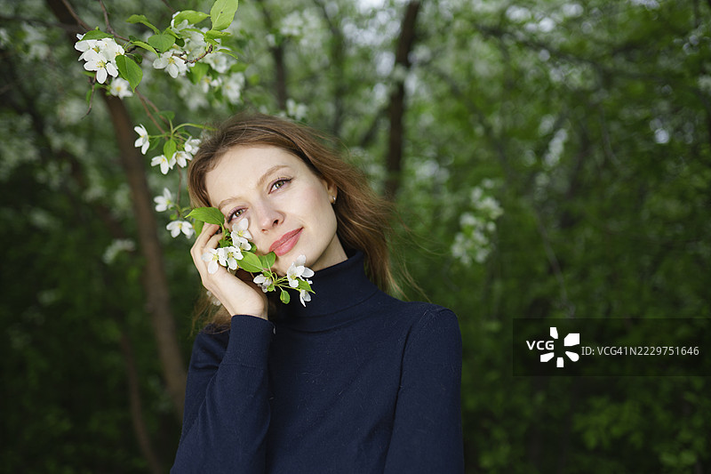 穿着高领毛衣的女人在户外微笑，身旁是一棵盛开的苹果树图片素材