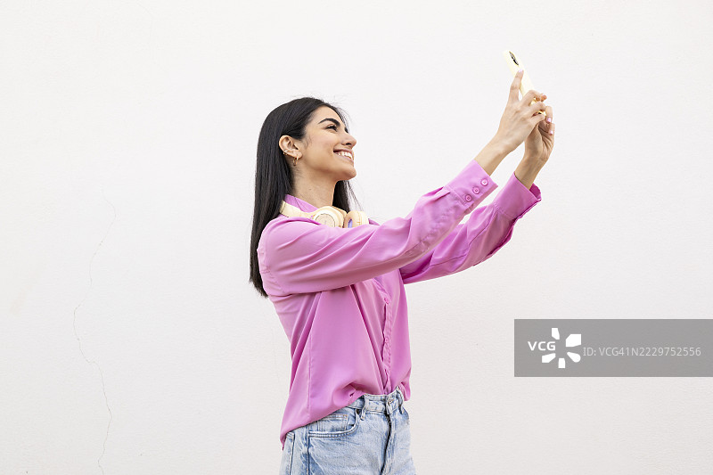 穿着粉色衬衫的女性在白色墙壁前用智能手机自拍图片素材