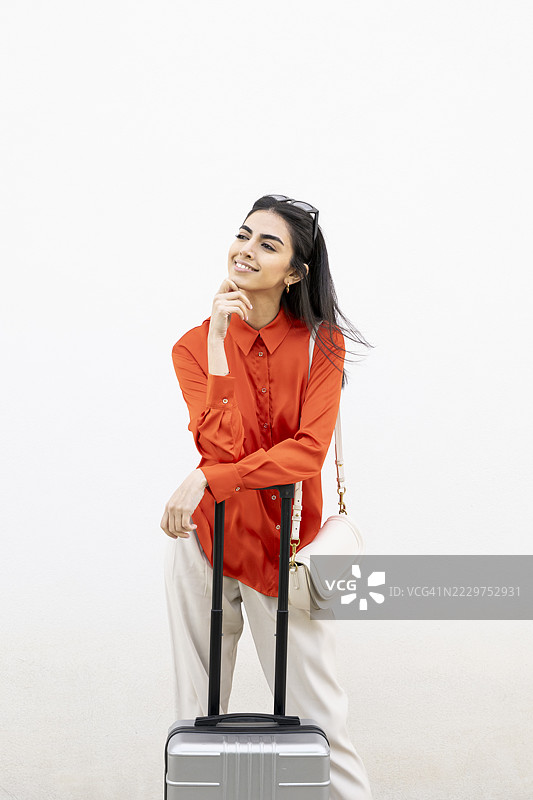 穿着橙色衬衫的女人站在白色墙壁旁，手里拿着行李箱。图片素材