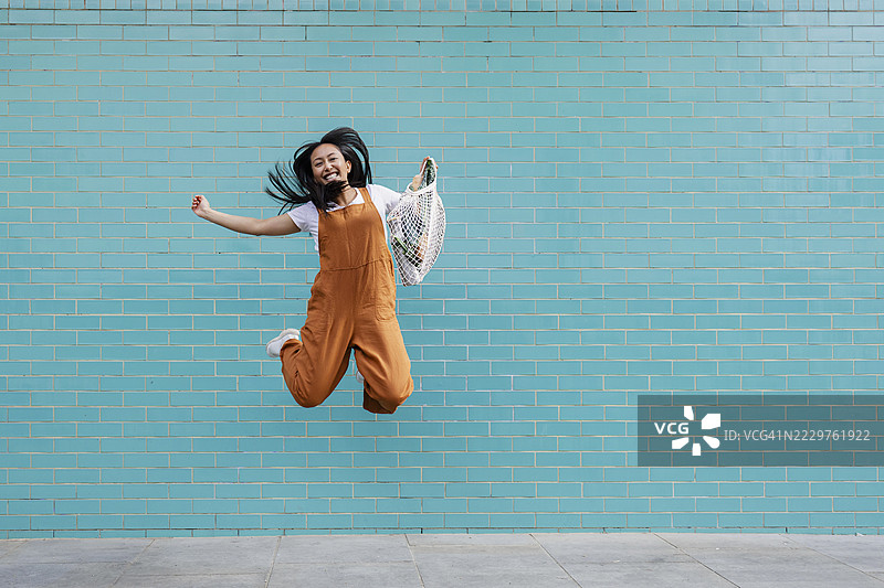 快乐的人在青绿色墙前跳跃，手里拿着可重复使用的购物袋图片素材