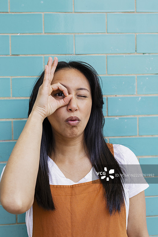 调皮的厨师穿着围裙，眨眼并做出“好”的手势，背景是青绿色的瓷砖墙。图片素材