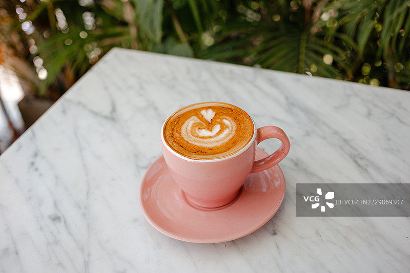 粉色杯子里的植物基牛奶卡布奇诺，放在大理石桌上。香气四溢的咖啡，配有美丽的拿铁艺术。图片素材