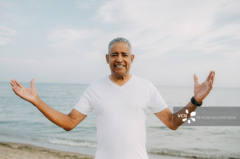 一位年长的男士站在沙滩上，开心地张开双臂，面带微笑。图片素材