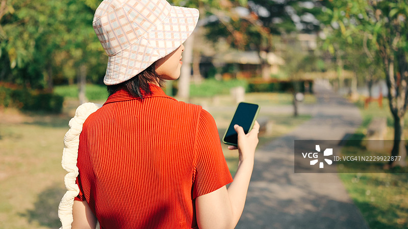 年轻的亚洲女性旅行者穿着时尚的红色连衣裙和宽边帽，在郁郁葱葱的公园户外使用手机，数字游牧者的夏季假期在亚太地区。图片素材
