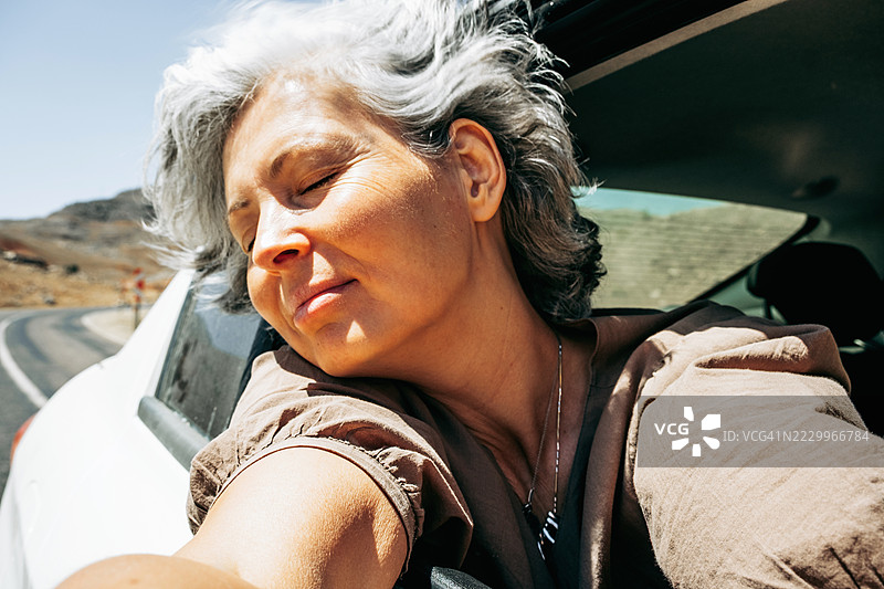 一位中年白人女性，灰发，身体探出车窗外，闭着眼睛，面带微笑。她的头发随风飘动，背景是干旱的风景。体现了宁静、正念时刻和独自自由的概念。图片素材