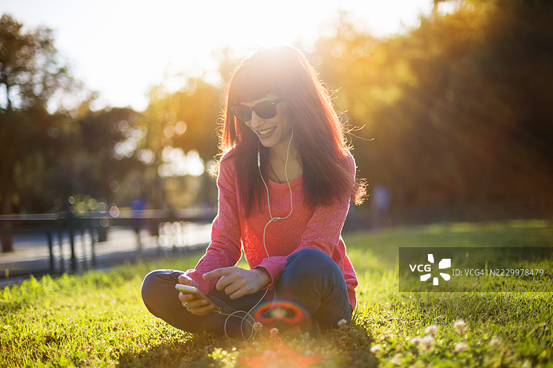 年轻女性在阳光明媚的公园里坐着，享受着手机音乐。图片素材