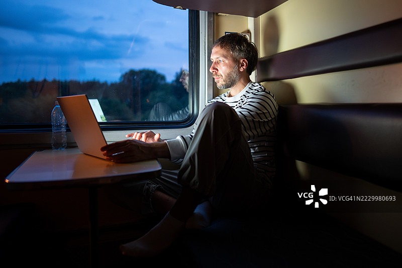 疲惫的男子在黄昏时分的火车车厢内使用笔记本电脑。过度劳累的员工在旅途中进行数字工作。图片素材