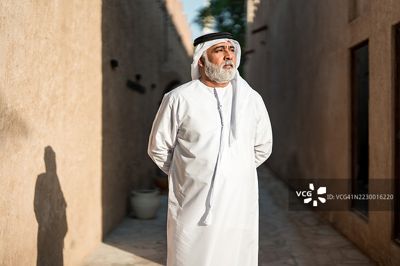 穿着传统白色坎杜拉的阿联酋老人在阳光明媚的小巷中漫步图片素材