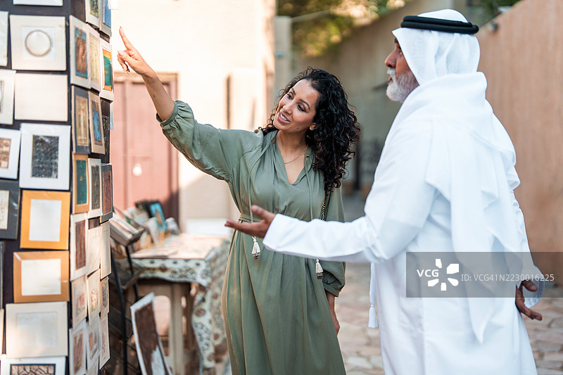 户外小店中，微笑的阿拉伯女性与年长男性老板互动，穿着传统服饰。图片素材
