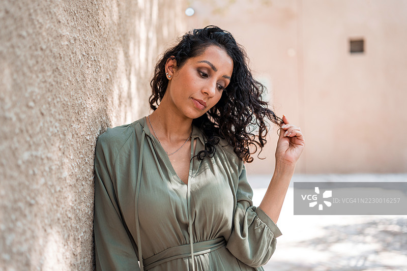 优雅的中年阿拉伯女性，卷发，穿着绿色连衣裙，倚靠在有纹理的墙上图片素材