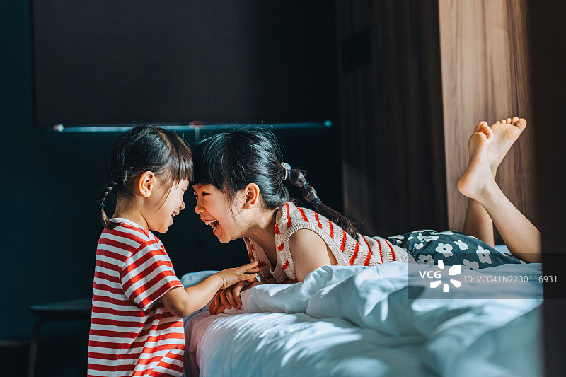 两个快乐的亚洲姐妹在温馨的家中床上玩耍，享受乐趣。女孩们面带微笑，互相拥抱。幸福的童年，无条件的姐妹情。兄弟姐妹之间的爱与亲密，家庭的爱与团聚。图片素材