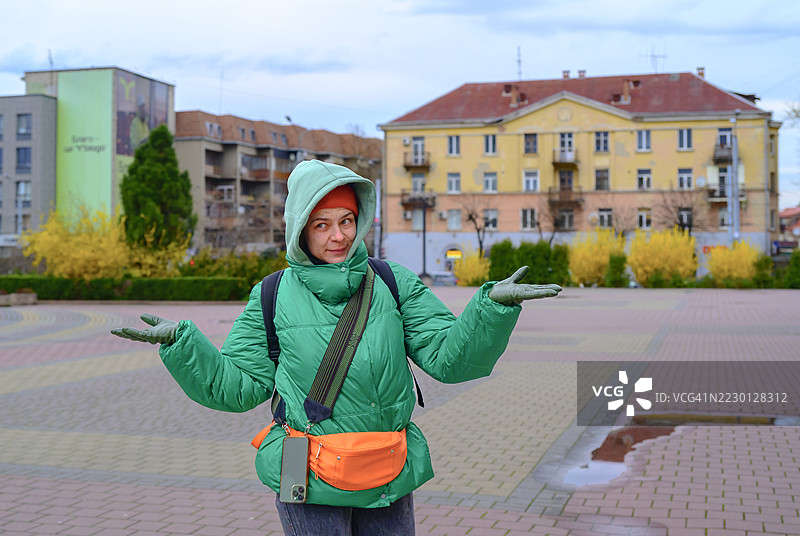 穿着绿色夹克和橙色腰包的女性旅行者在乌克兰阴云密布的天空下，犹豫地探索伊万诺-弗兰基夫斯克的鹅卵石街道。图片素材