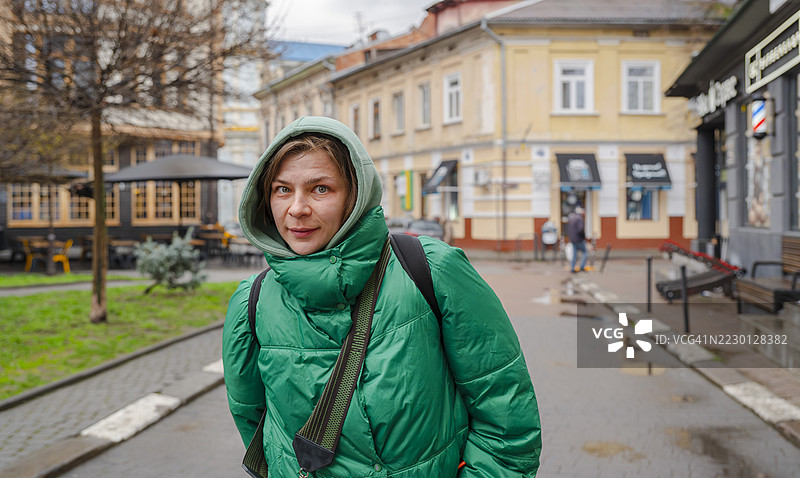 中年女性穿着绿色连帽夹克，站在繁忙的城市街道上，平静地凝视着镜头，展现了乌克兰利沃夫的放松城市生活方式。图片素材