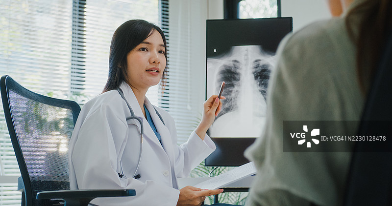 年轻的亚洲女性医生在现代医疗诊所办公室分析肺部X光片，并向患者解释诊断。图片素材