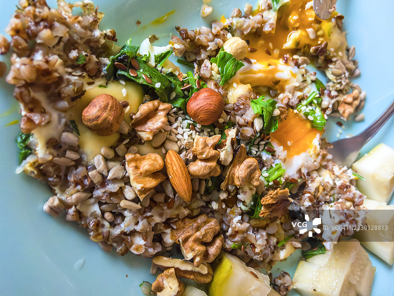 木桌上放着蓝色陶瓷盘，盘中是用金色蛋黄、散落的坚果和香蕉片装饰的荞麦粥，构成了一顿营养丰富的早餐。图片素材