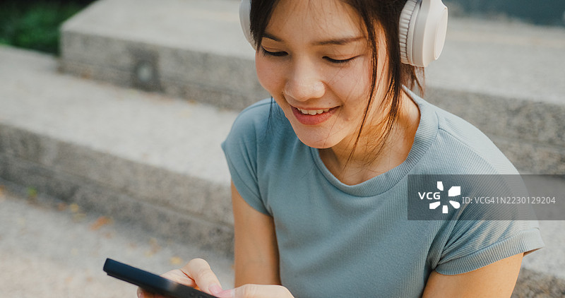 快乐的年轻亚洲女性在公园户外放松，戴着耳机，用智能手机听音乐。图片素材