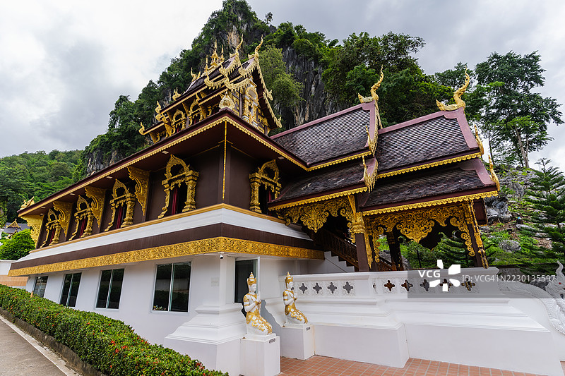 一座装饰华丽的泰国寺庙，拥有精美的金色装饰和层叠的屋顶，矗立在郁郁葱葱的绿色山脉下，天空布满云彩。图片素材