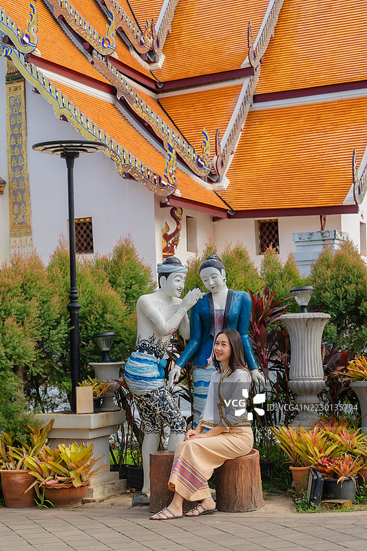 一位身穿传统泰国服饰的女性坐在寺庙花园中，旁边是色彩斑斓的低语雕像，背景是华丽的橙色屋顶。图片素材
