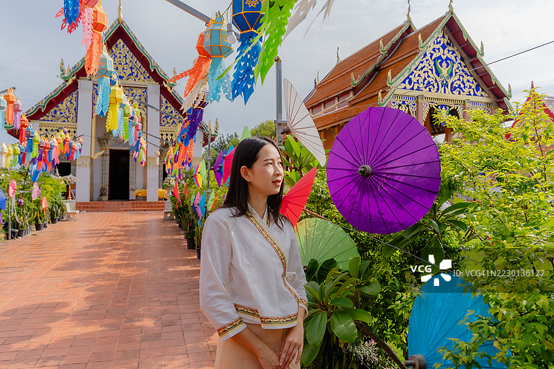 一位身穿传统泰国服装的女性站在装饰着五彩灯笼和伞的热闹寺庙走廊中，庆祝节日。图片素材
