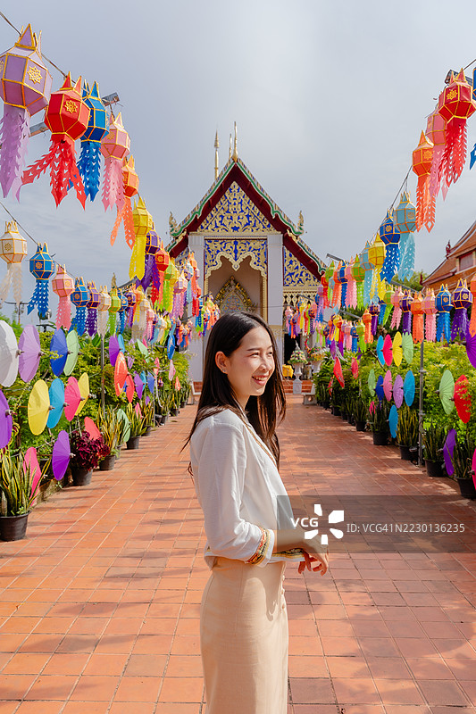一位身穿传统泰国服装的女性站在装饰着五彩灯笼和伞的热闹寺庙走廊中，庆祝节日。图片素材