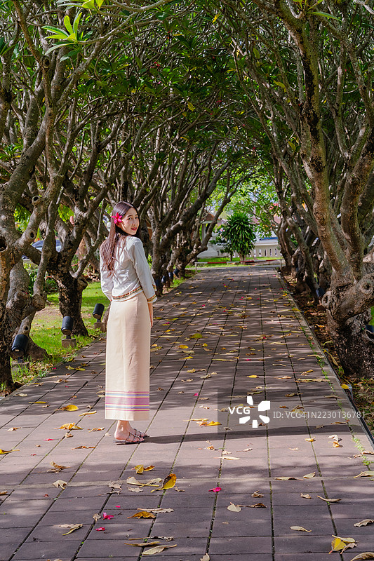 一位身穿传统泰国服饰的女性在郁郁葱葱的树木拱形隧道下，耳边夹着一朵粉色花朵。图片素材