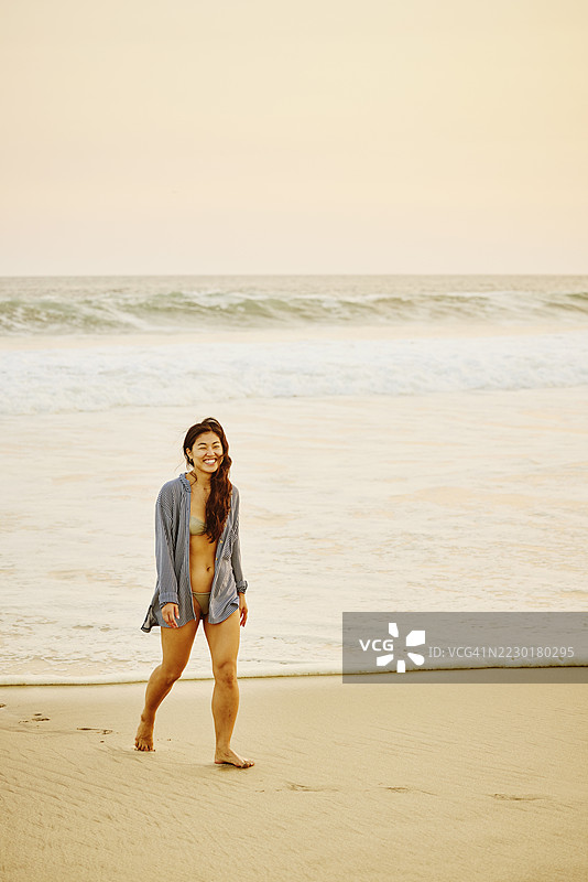 在日落时分，穿着泳装的微笑女性在热带海滩上漫步的广角镜头图片素材