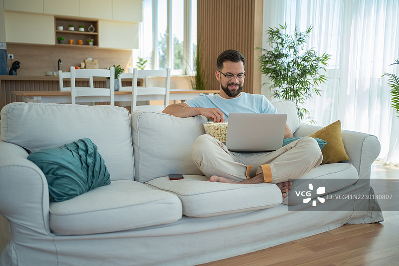 年轻男子坐在沙发上放松，观看笔记本电脑上的电影，吃着爆米花图片素材