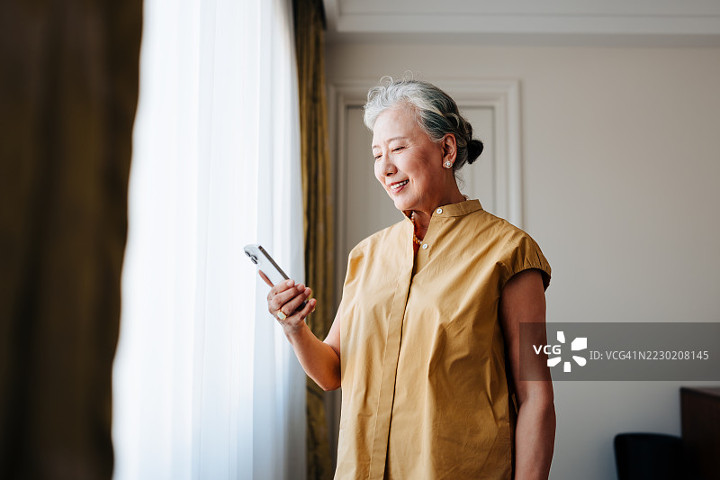 亚洲老年女性在家中窗边放松时使用智能手机。图片素材