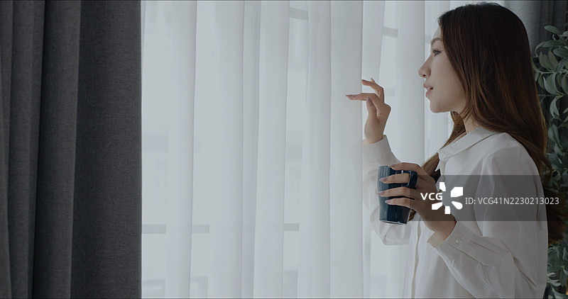 微笑的亚洲女性在宁静的家中窗边享受温暖的饮品图片素材