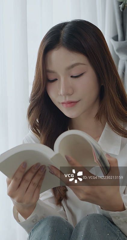 亚洲女性在明亮舒适的房间窗边阅读一本书，体现了正念、教育、放松、个人发展和宁静的家庭生活方式。图片素材