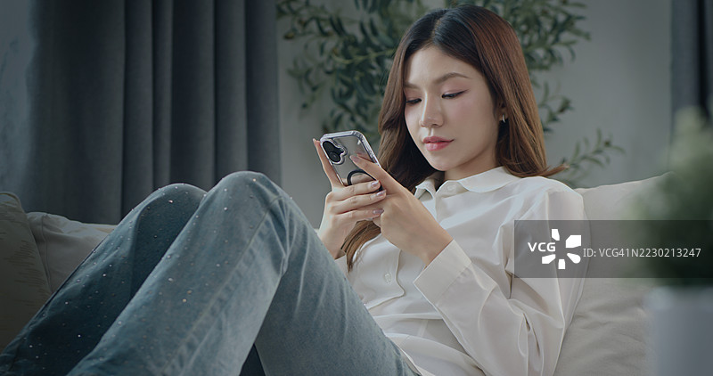 年轻的亚洲女性在家中沙发上放松，使用智能手机，代表数字生活方式、社交媒体、在线沟通、移动技术和休闲生活。图片素材