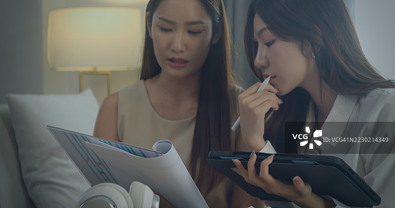 两位年轻的亚洲女性在家中讨论文件和图表，展现了团队合作、协作和专注沟通的氛围，营造出一个休闲现代的工作空间。图片素材