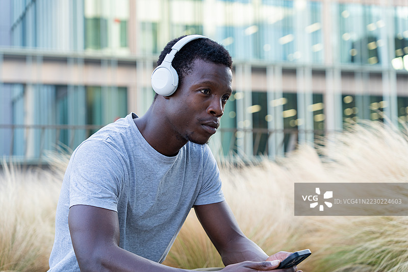 年轻的非裔美国男性在城市公园里戴着耳机听音乐，手里拿着智能手机图片素材