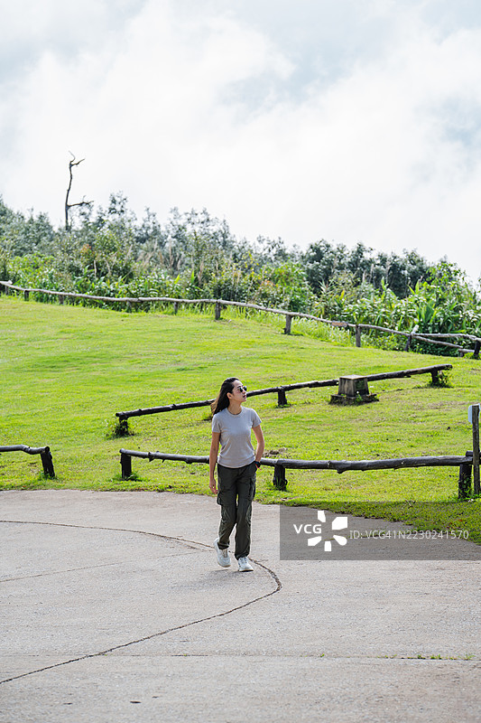 一位穿着休闲服的女性站在铺砌的山间小径上，四周环绕着郁郁葱葱的绿树，俯瞰着云雾缭绕、宁静的背景。图片素材