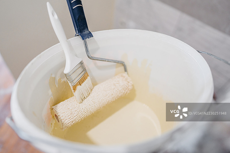 一个油漆滚筒和一把油漆刷放在一个油漆桶里，位于一个正在装修的空公寓内图片素材