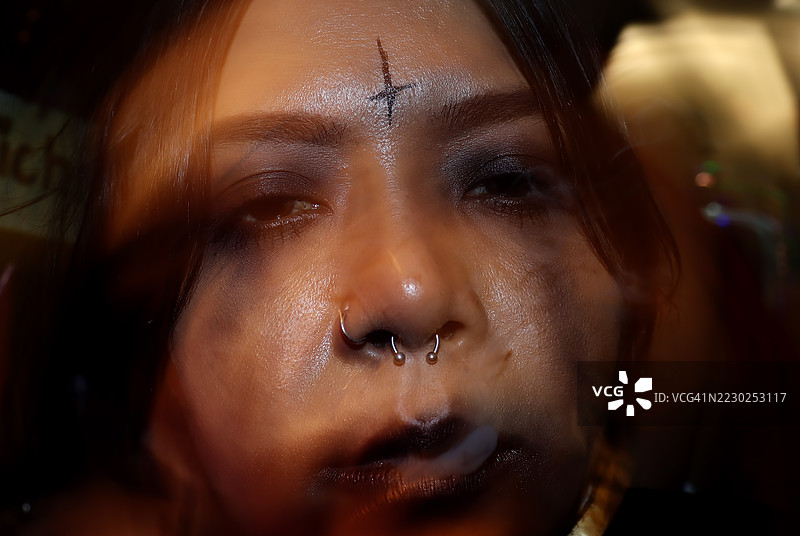 特写肖像，展示一位个性张扬的人，鼻中隔穿孔，额头上有黑色倒十字，吐出烟雾，充满戏剧性。万圣节派对化妆概念。图片素材