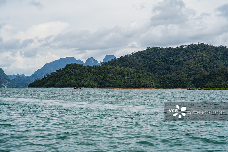 一个郁郁葱葱、树木繁茂的岛屿坐落在碧绿色的湖中央，四周环绕着遥远的石灰岩山脉，天空布满云彩。图片素材