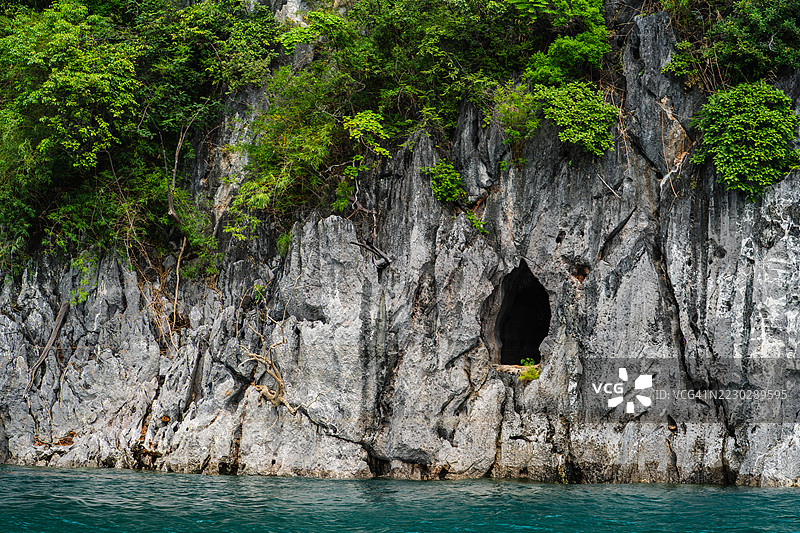 一座崎岖的石灰岩悬崖上有一个黑暗的洞口，耸立在碧绿色的湖水中，周围环绕着郁郁葱葱的热带植被。图片素材