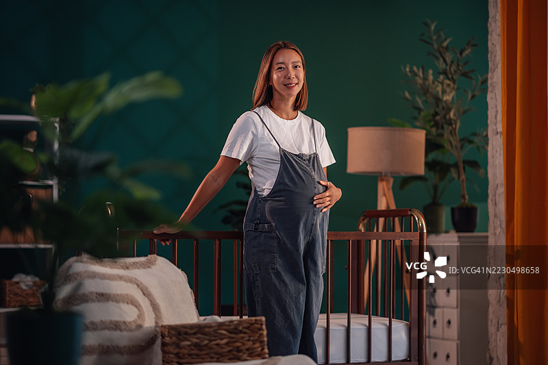 孕妇轻柔地抚摸肚子，站在婴儿床旁边图片素材