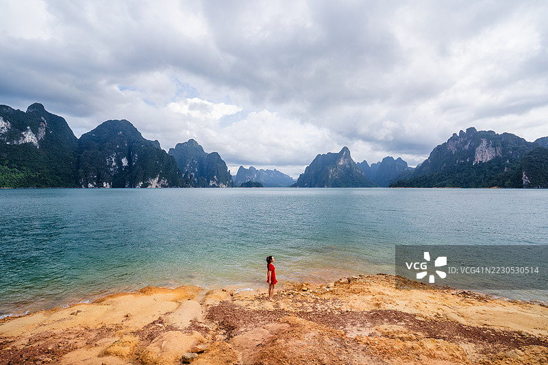 一位穿着红色裙子的女性独自站在碧绿色湖边，凝视着阴云密布的天空下壮观的石灰岩悬崖。图片素材