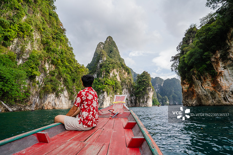 一名穿着花衬衫的男子坐在木船上，平静地欣赏着热带环境中翡翠般的水域和高耸的石灰岩悬崖。图片素材