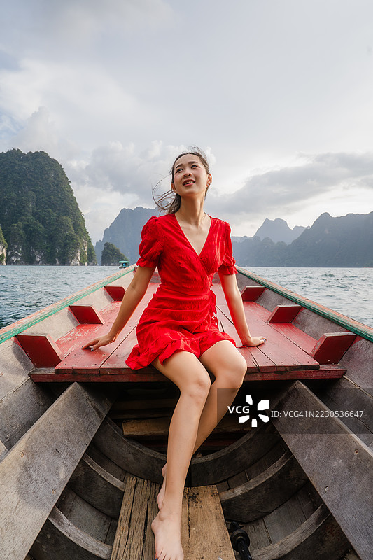 一位穿着红色连衣裙的女性赤脚坐在木船上，享受着被群山环绕的美丽湖泊，正值黄金时刻。图片素材