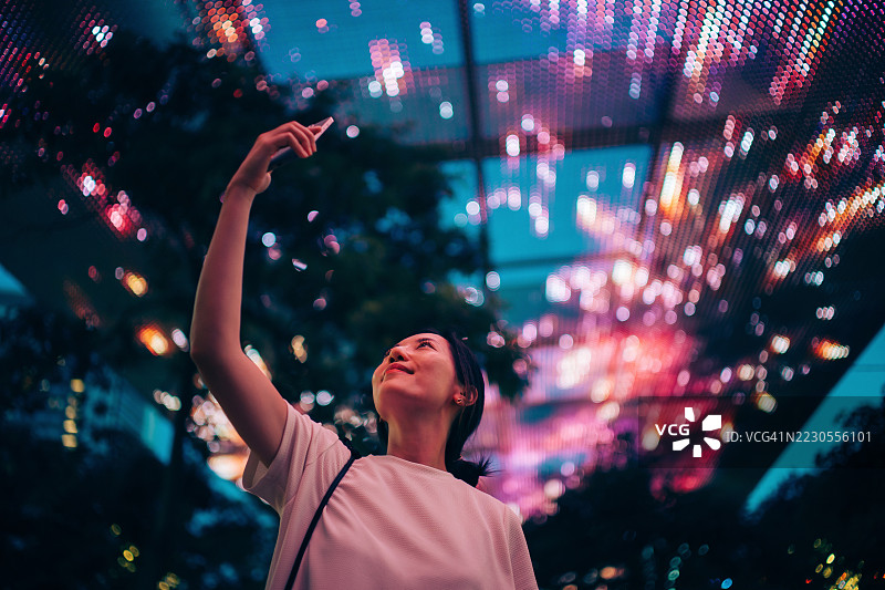 微笑的年轻亚洲女性在城市街道的步行桥下拍摄照明的LED灯饰。虚拟现实和增强现实技术的概念。元宇宙和未来数据。虚拟助手。人们、人工智能、创新和技术。图片素材