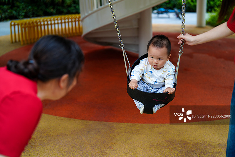 亚洲小男孩在秋千上，祖母俯身看着宝宝，他们互相凝视。这个一岁的小宝宝还不会走。图片素材