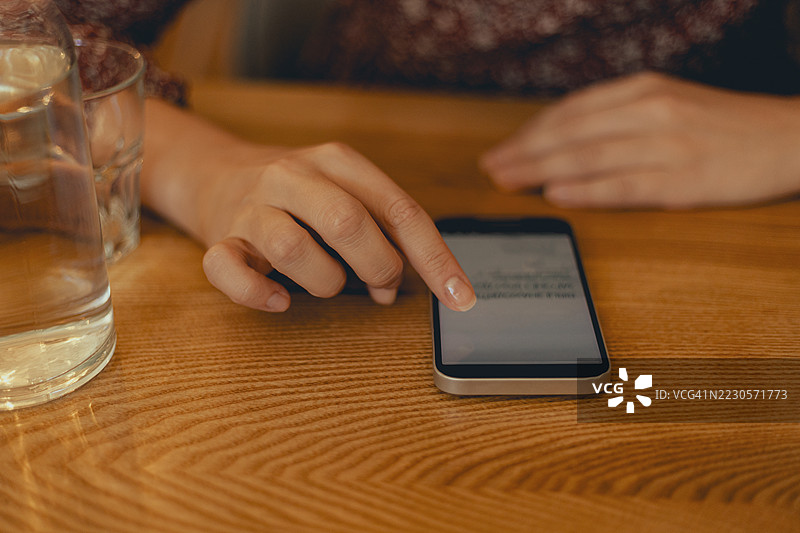 年轻女性在木桌上使用智能手机，旁边有一瓶饮料和一个玻璃杯。图片素材