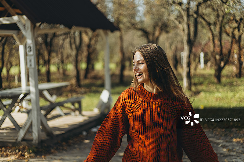 年轻女性在阳光明媚的秋季花园中靠在凉亭旁，开心地笑着。图片素材