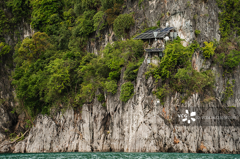 一座小房子装有太阳能电池板，依附在崎岖的石灰岩悬崖上，四周被浓密的热带森林环绕，下面是碧蓝的海水。图片素材