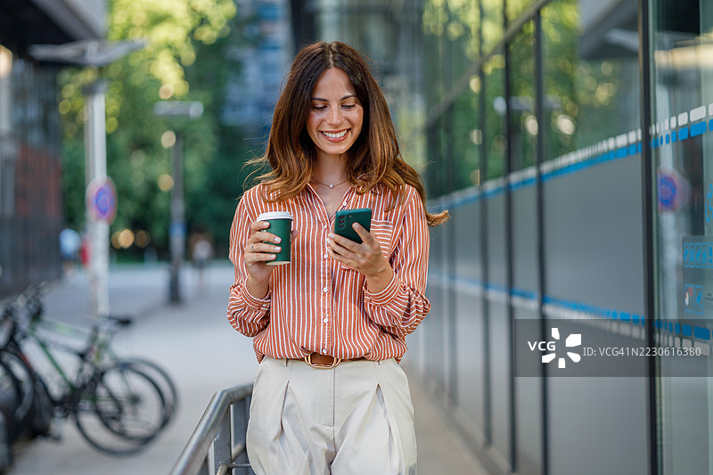 年轻女商人在阳光明媚的日子里，走在充满活力的城市街道上，一边享受咖啡，一边使用智能手机。图片素材